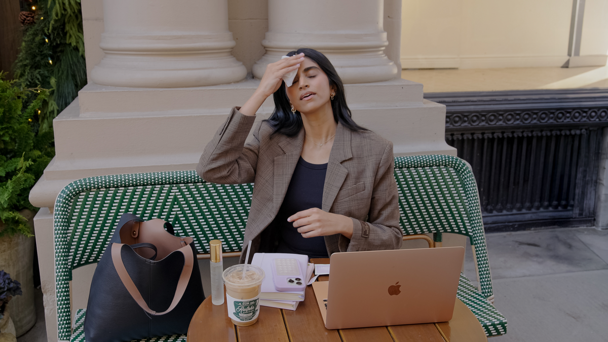 Woman sitting at a table with a laptop and coffee, looking stressed outdoors.