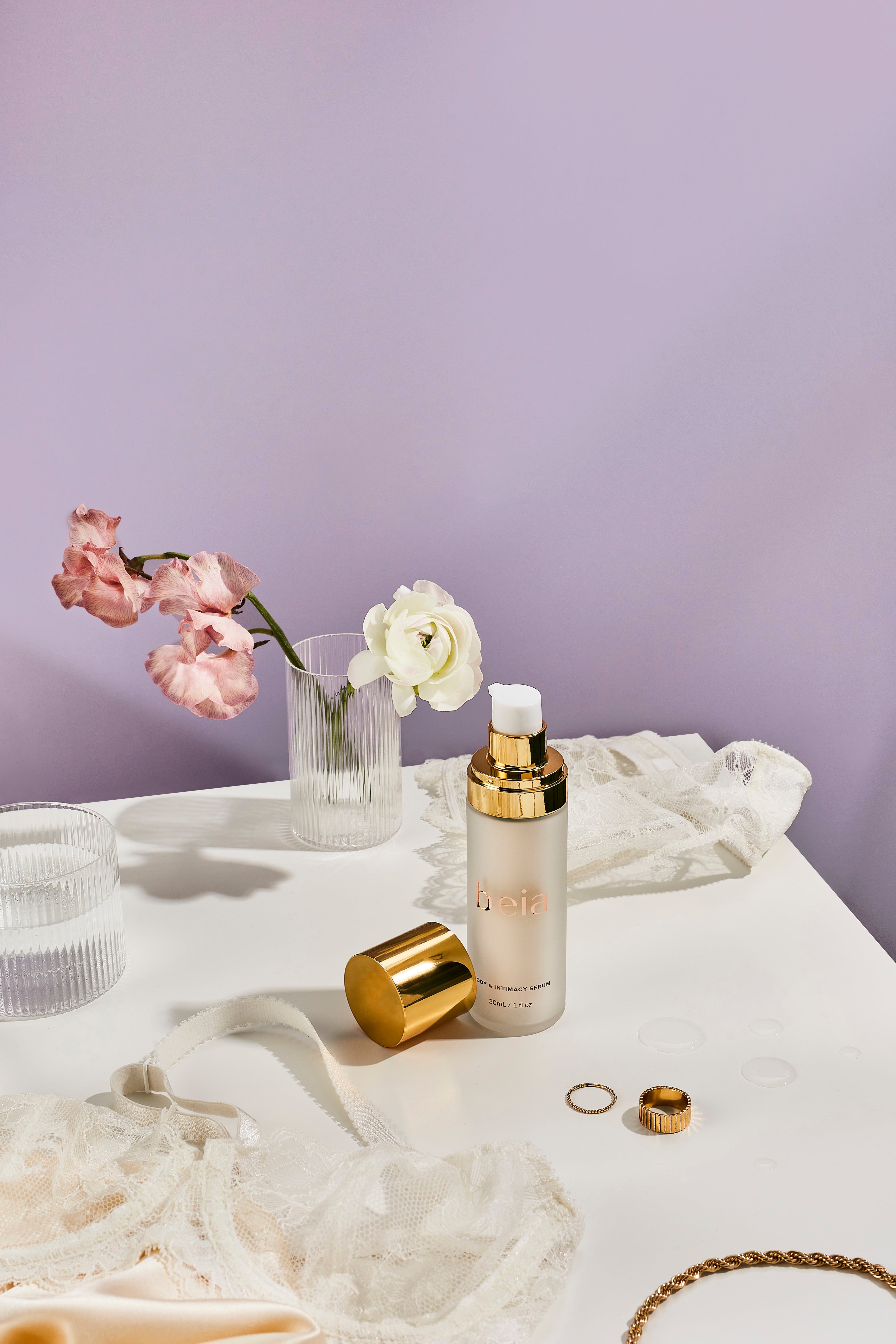 Skincare serum bottle with gold cap on a purple background with flowers and jewelry 